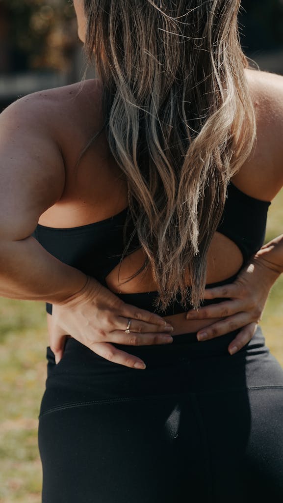 A woman in activewear stretching her back outdoors in a sunlit park setting.